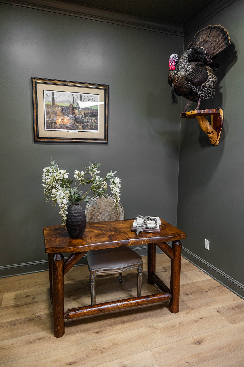 May include: A wooden desk with a live-edge top and log-style legs, paired with a cane-back chair. On the desk sits a black textured vase with white floral stems, a stack of two books, and a beaded strand. A taxidermy turkey is mounted on the wall.