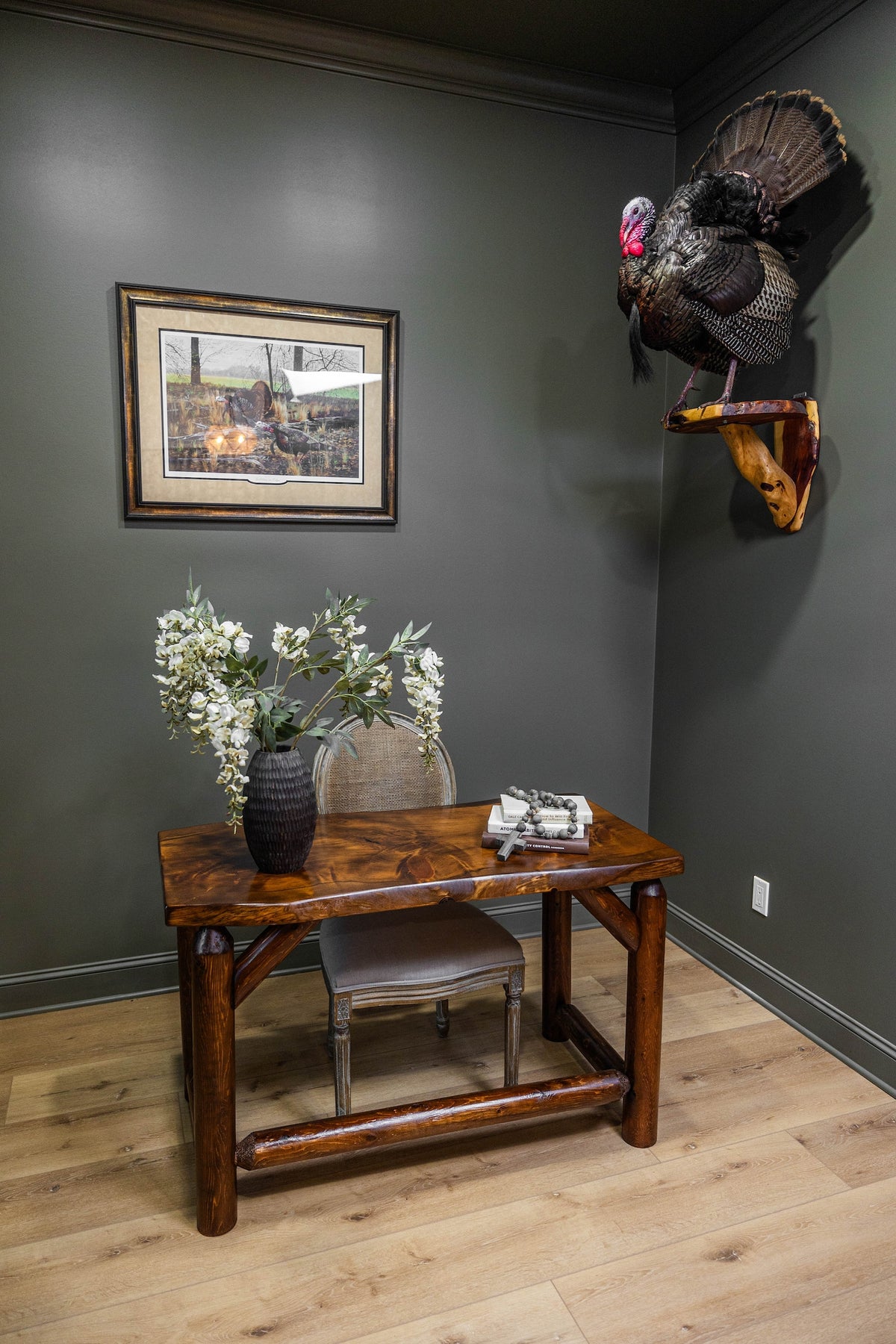 May include: A wooden desk with a live-edge top and log-style legs, paired with a cane-back chair. On the desk sits a black textured vase with white floral stems, a stack of two books, and a beaded strand. A taxidermy turkey is mounted on the wall.