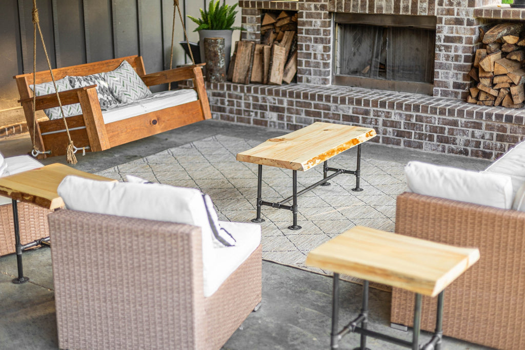 May include: A patio furniture set with a wooden swing, two wicker chairs, and three wooden tables with black metal legs. The furniture is arranged around a brick fireplace with a wood-burning stove.