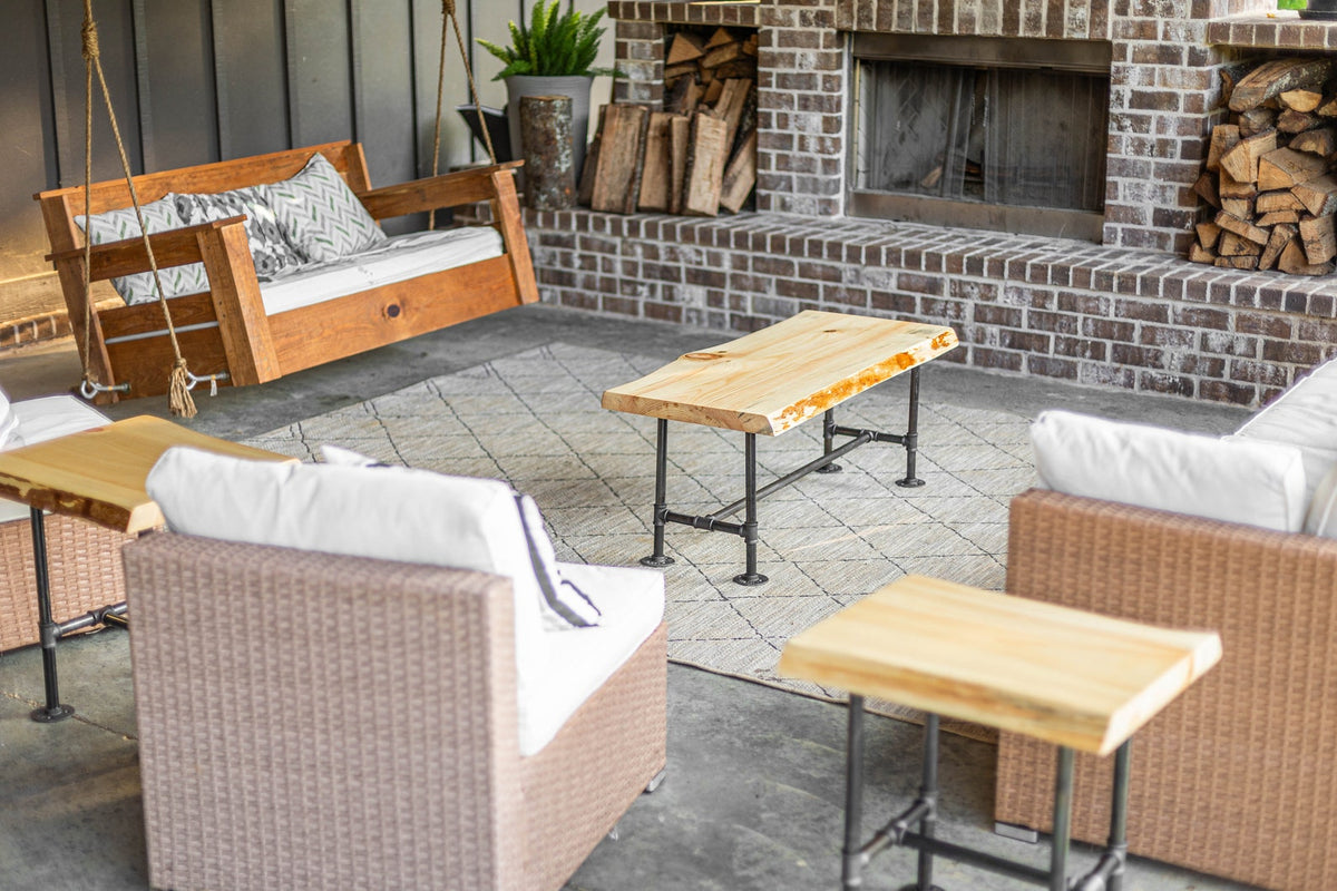 May include: A patio furniture set with a wooden swing, two wicker chairs, and three wooden tables with black metal legs. The furniture is arranged around a brick fireplace with a wood-burning stove.