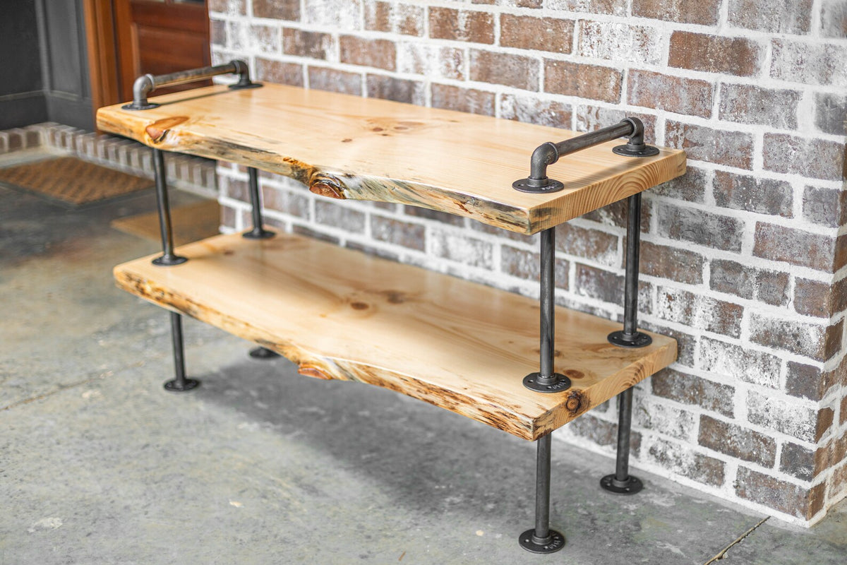 May include: A two-tiered wooden shelf with a live edge design, supported by black metal pipes. The shelf is made of light wood and has a natural finish. The shelf is mounted to a brick wall.