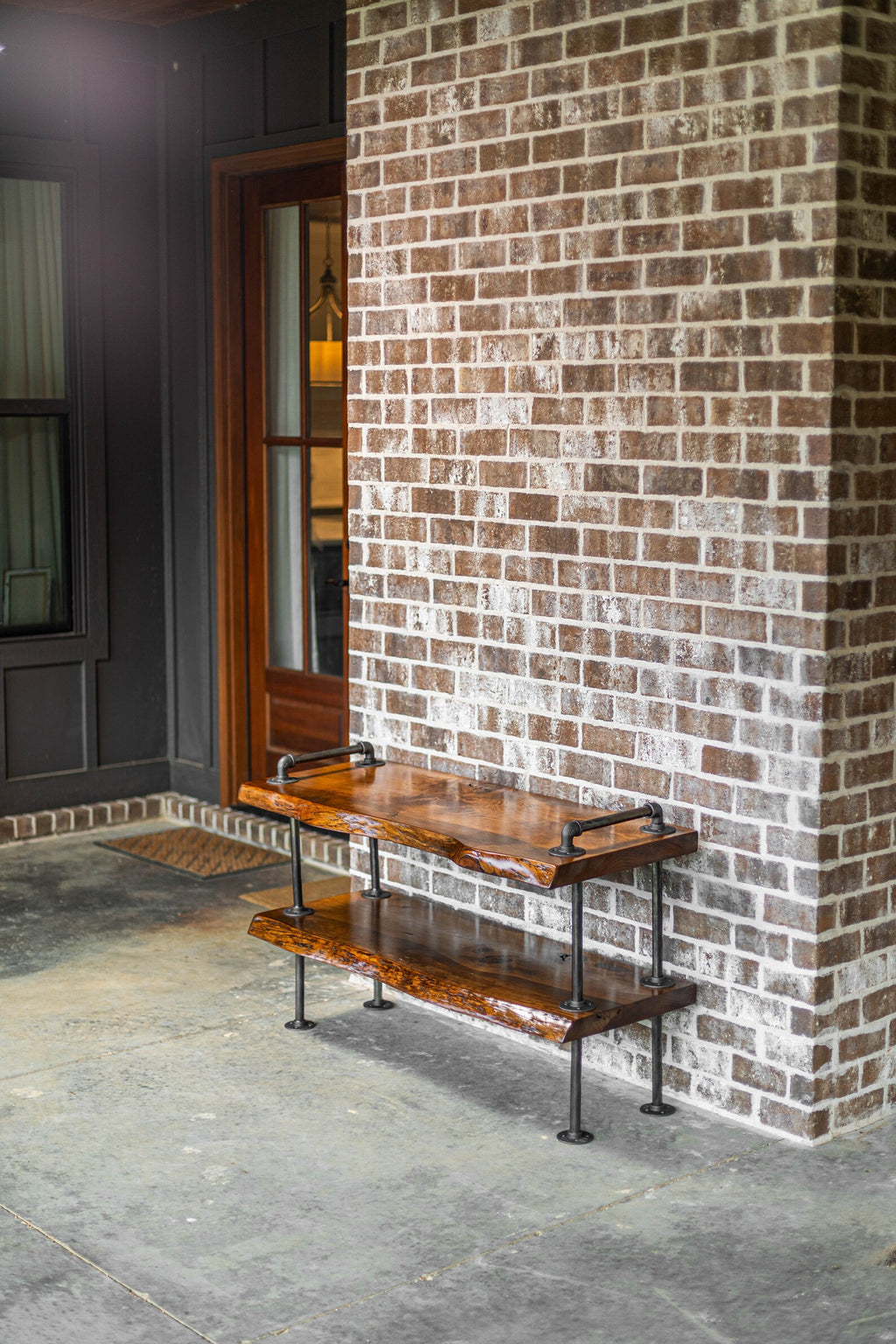 May include: A two-tiered wooden shelf with black metal pipe legs. The shelf is made of natural wood with a rustic finish. The shelf is sitting on a concrete patio in front of a brick wall.