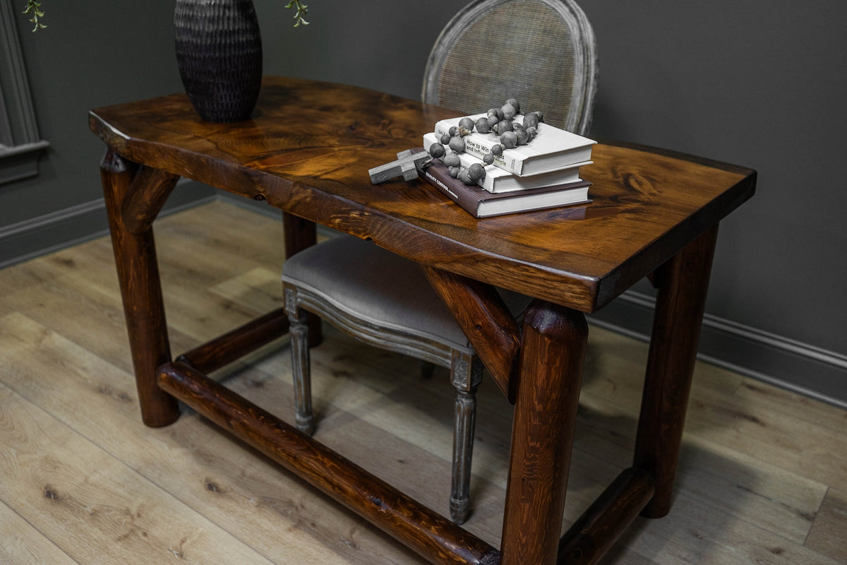 May include: A rectangular wooden desk with a dark brown finish and visible wood grain, featuring thick, sturdy legs and a lower support frame. On the surface sits a stack of two books, a string of dark grey beads, and a small wooden cross.