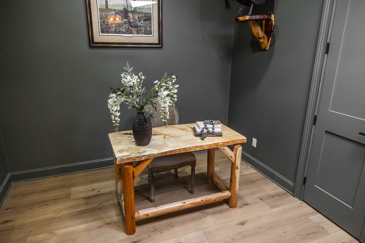 May include: A rectangular desk with a light-colored natural wood slab top and thick, cylindrical log legs. A dark textured vase filled with white floral branches sits on the surface alongside a stack of books and a decorative chain link object.