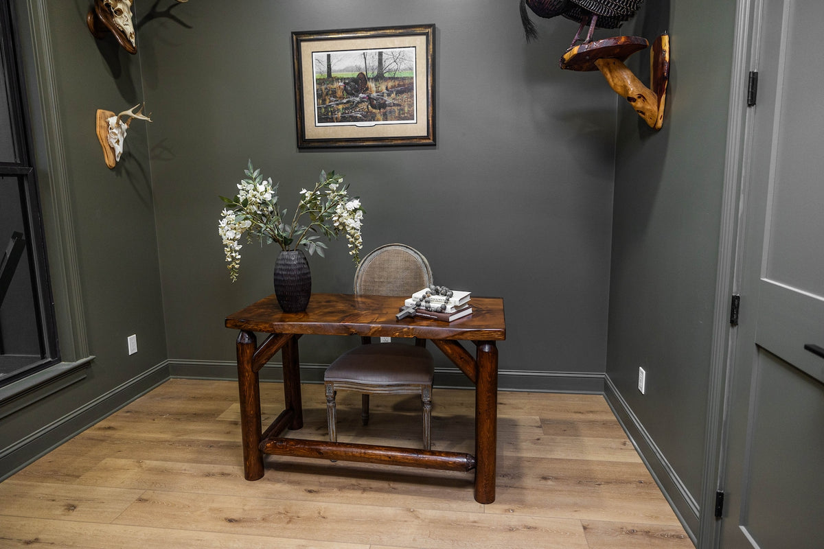 May include: A dark brown wooden desk with a thick, polished rectangular top and sturdy legs connected by horizontal support beams. The desk is positioned in a room with dark green walls, featuring a gray upholstered chair, a black vase with white flowers, and a stack of books on the surface.