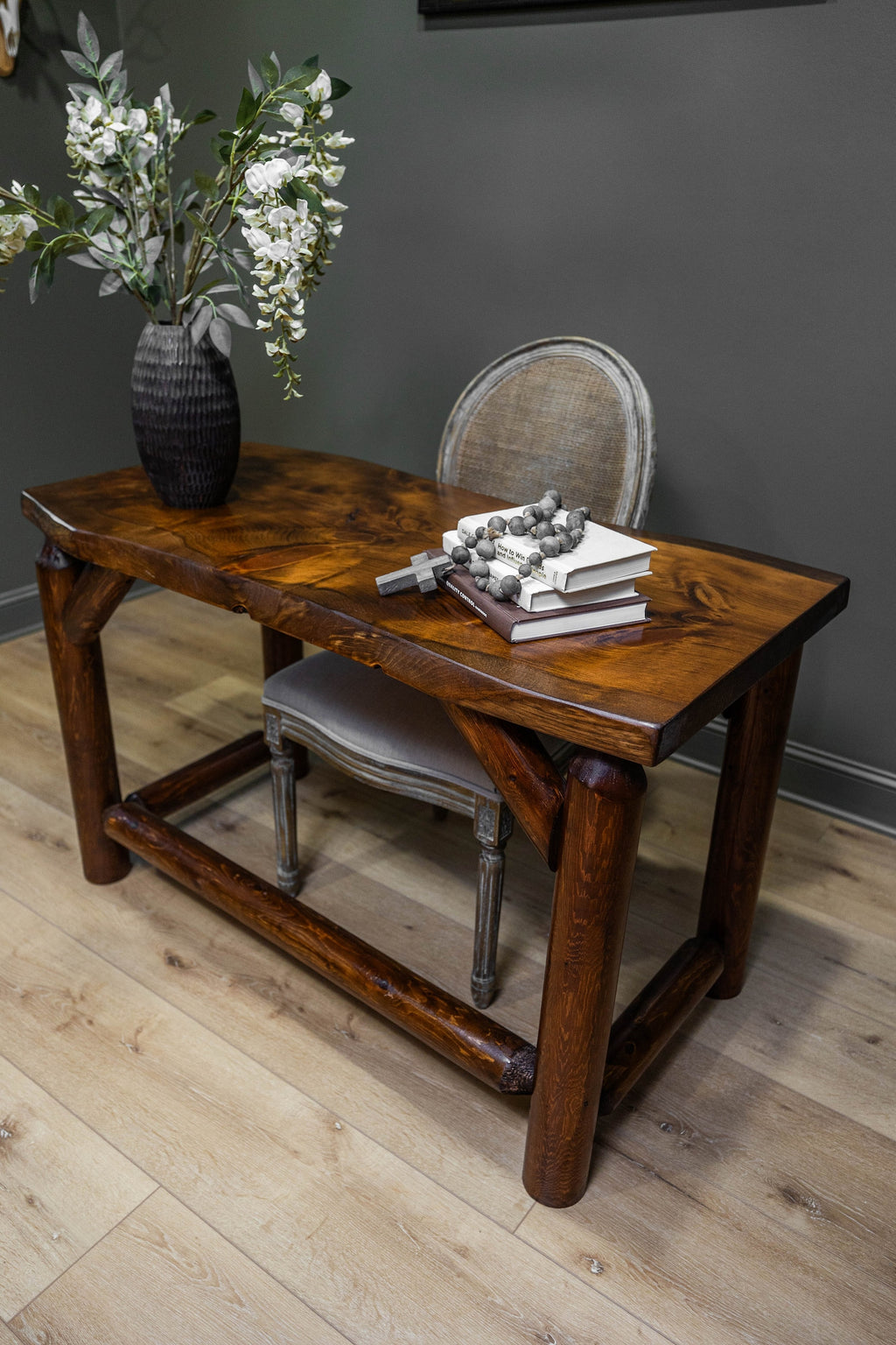 May include: A dark brown wooden desk with a thick, natural-edge top and sturdy cylindrical legs connected by a lower support beam. On the desk sits a dark textured vase with white flowers, a stack of two books, a wooden cross, and a beaded garland.