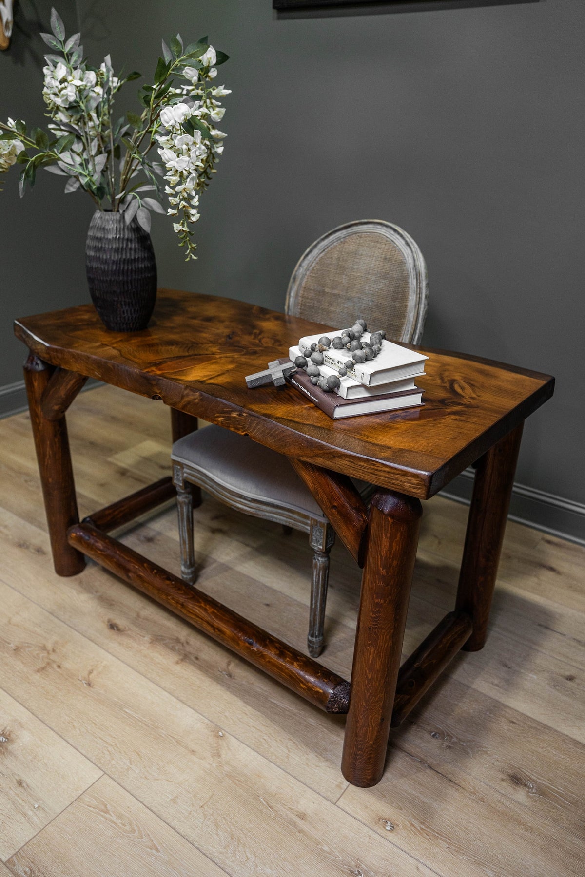 May include: A dark brown wooden desk with a thick, natural-edge top and sturdy cylindrical legs connected by a lower support beam. On the desk sits a dark textured vase with white flowers, a stack of two books, a wooden cross, and a beaded garland.