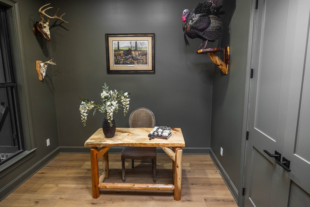 May include: A live-edge wooden desk with a natural grain top and sturdy log legs, paired with a gray upholstered chair. On the desk sits a dark vase with white floral stems and a stack of books. The room features dark walls, taxidermy, and art.
