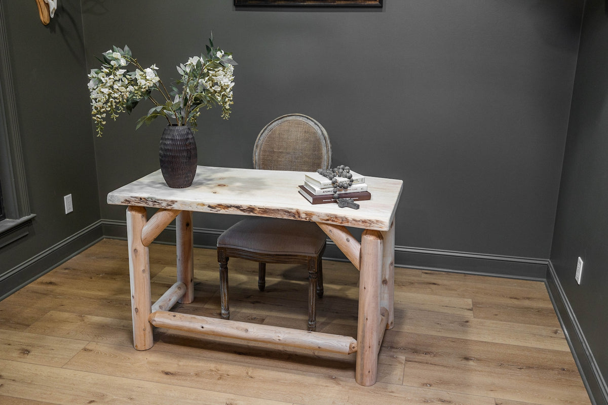 May include: A rectangular desk featuring a light-colored natural wood slab top with live edges, supported by four thick, cylindrical log legs and a lower horizontal log stretcher. A beige upholstered chair is positioned behind the desk.