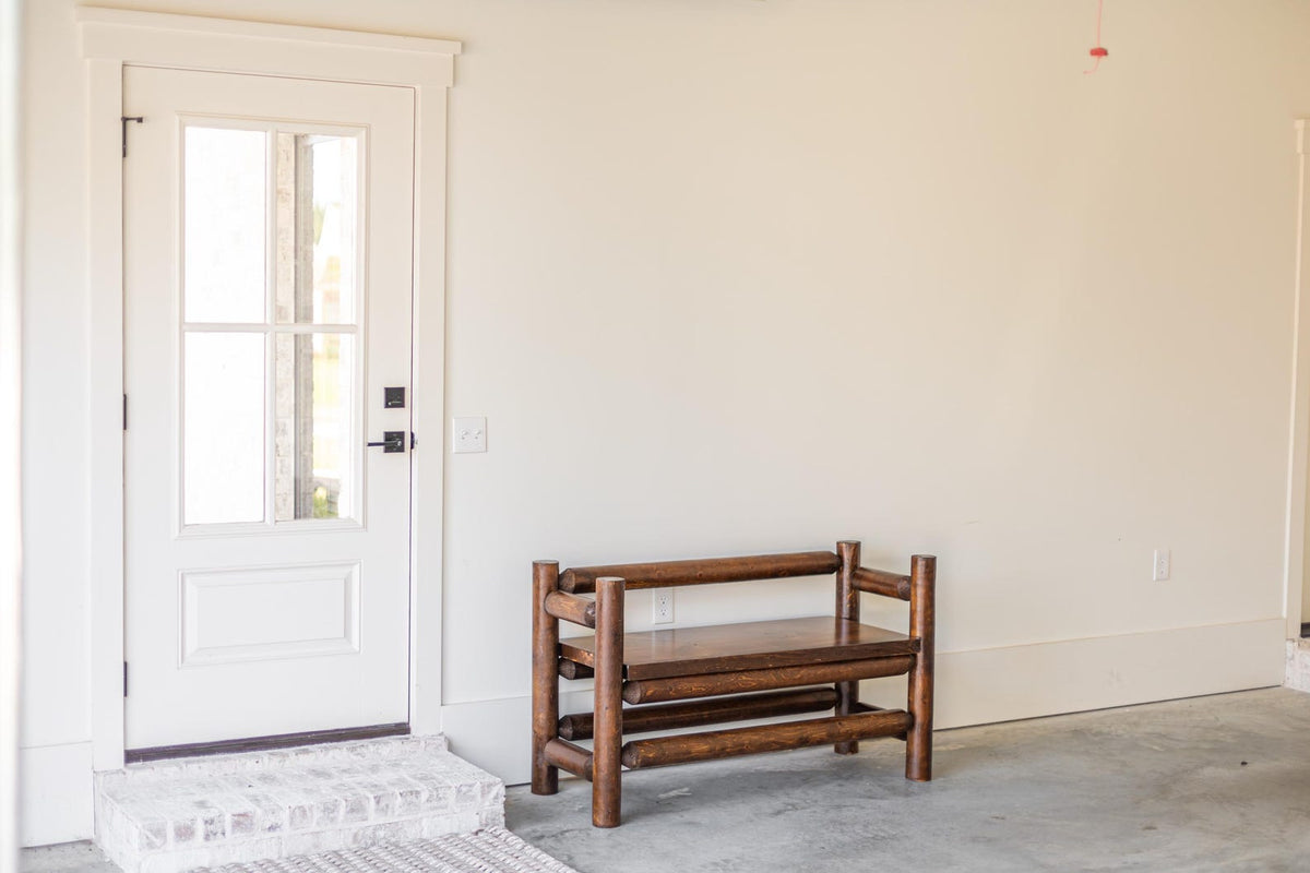May include: A dark brown wooden bench constructed from cylindrical logs with a flat seat and a backrest, positioned against a plain off-white wall next to a white door featuring a glass window pane and a black handle.