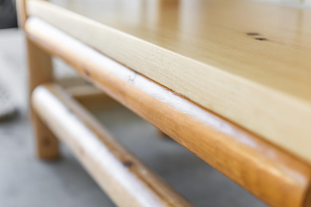 May include: A light-colored wooden bench featuring a smooth, flat top surface and rounded, cylindrical support beams underneath. The wood grain is visible, showcasing a natural finish with variations in tone across the structure.