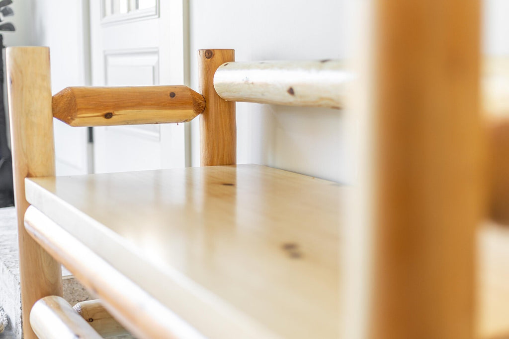 May include: A light-colored wooden bench constructed from smooth, polished logs. The bench features vertical log supports, a flat seat, and a horizontal log backrest with a faceted, hexagonal-shaped wooden accent piece on the side frame.