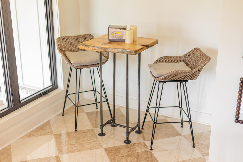 May include: A wooden table with a live edge top and black metal legs. Two wicker chairs with beige cushions are placed around the table. The table is decorated with a small box and two candles.