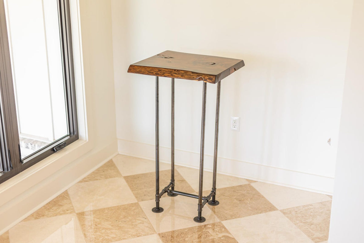 May include: A rustic wooden table top with live edge details sits on a black metal pipe stand with four legs. The table is in a room with white walls and a tiled floor.