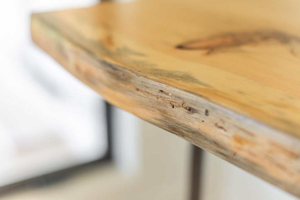 May include: Close-up of a light wood table top with a live edge. The wood has a natural, rustic look with a light brown color and a slightly darker brown edge.