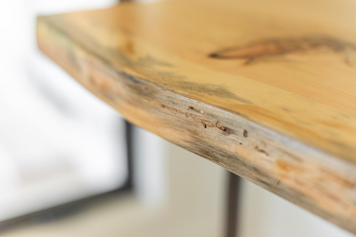 May include: Close-up of a light wood table top with a live edge. The wood has a natural, rustic look with a light brown color and a slightly darker brown edge.