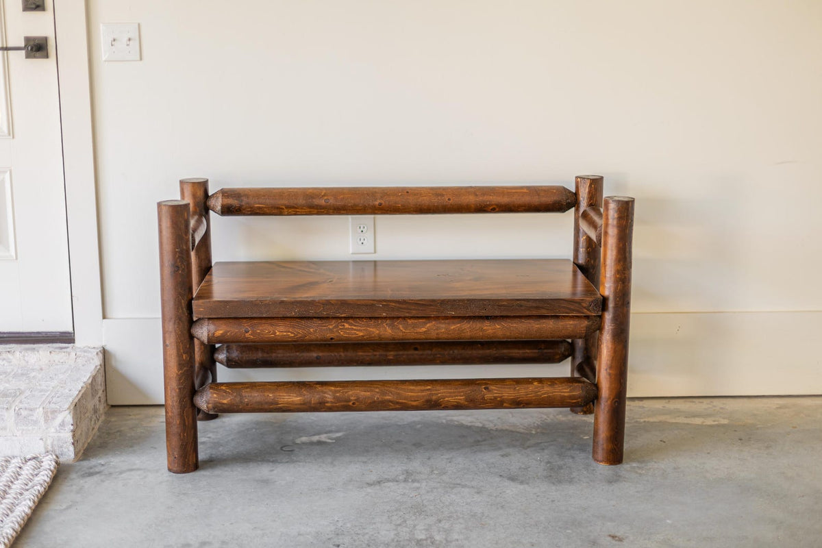 May include: A dark brown wooden bench constructed from cylindrical logs. The bench features a flat rectangular seat, a single horizontal log backrest, and four vertical log legs connected by horizontal log supports at the base and sides.