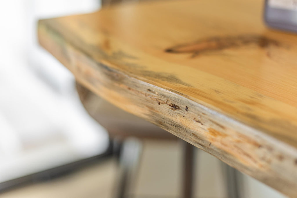 May include: Close-up of a light wood table top with a live edge. The edge of the table is a natural, irregular shape with a light brown and gray color.
