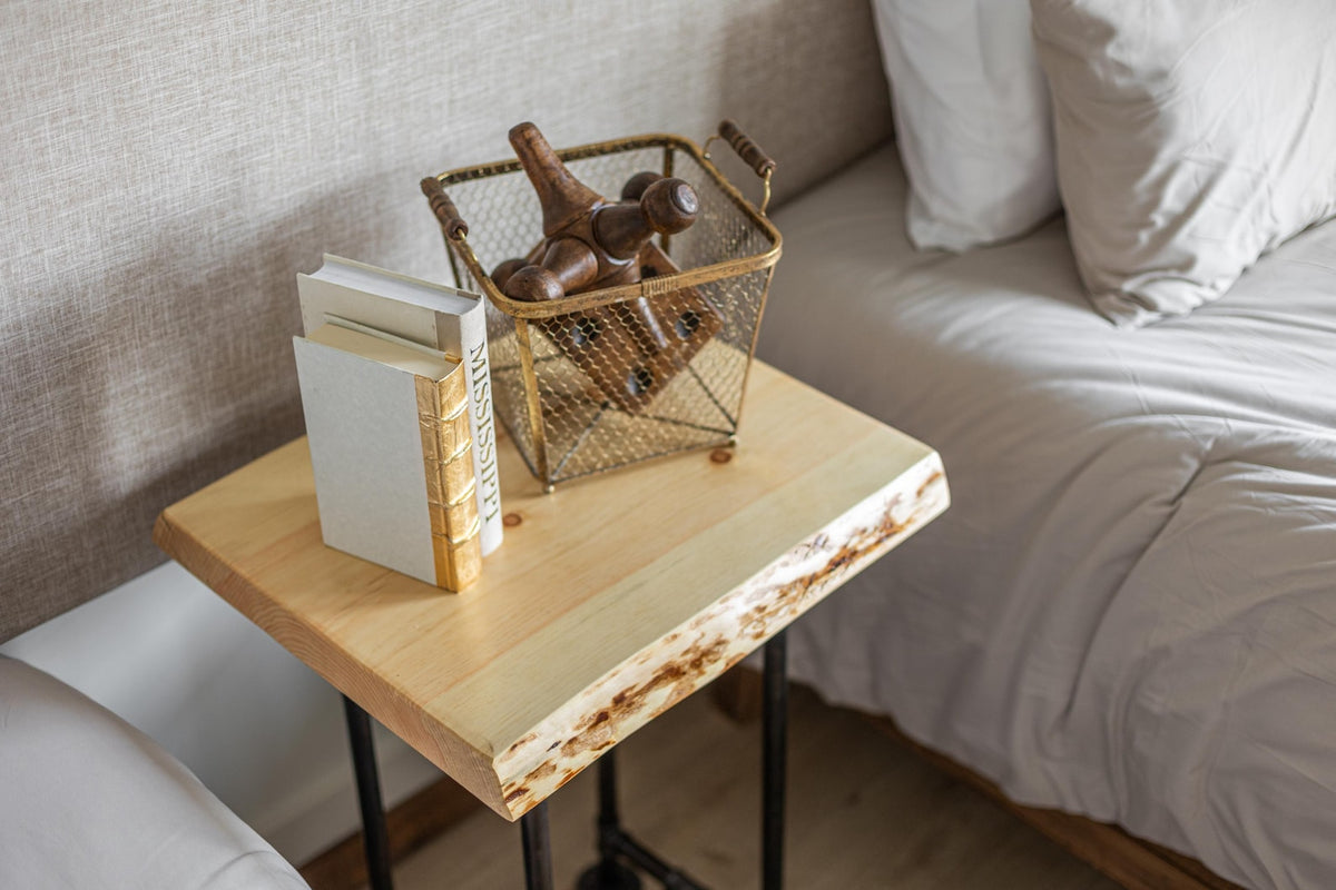 May include: A wooden side table with a live edge top and black metal legs. A white book with a gold spine and a wire basket filled with wooden objects are on the table.
