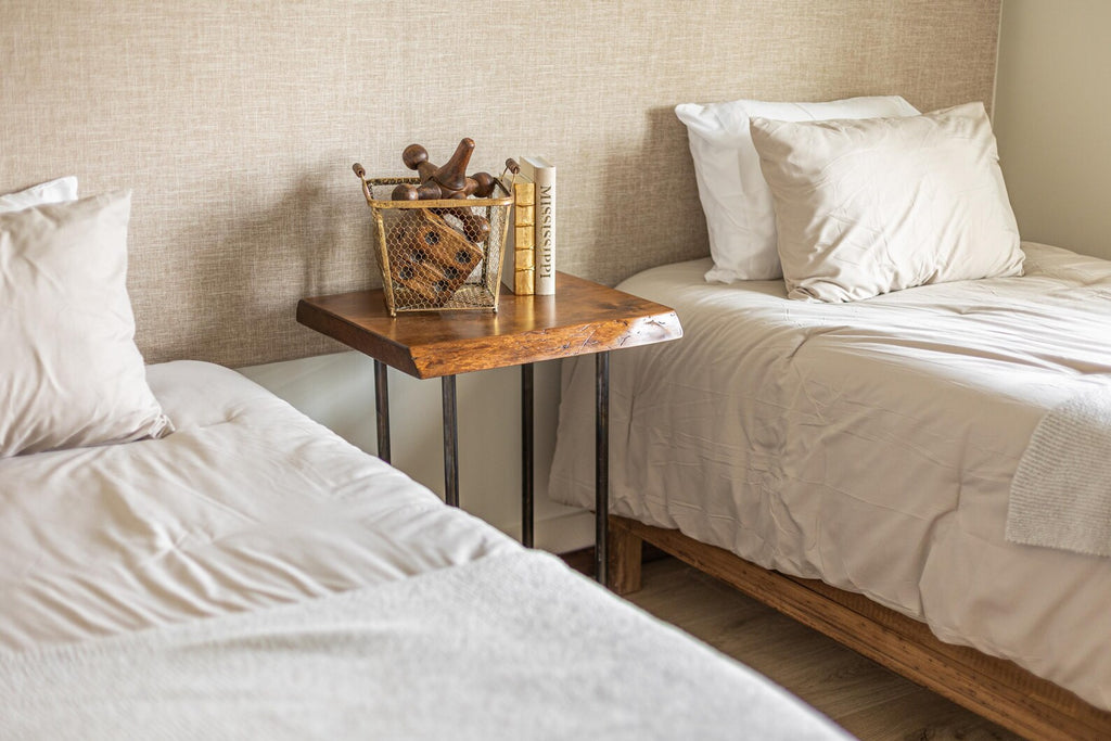 May include: Two twin beds with white linens and pillows, a wooden nightstand with a metal frame, a wire basket with wooden decorative objects, and two books on top of the nightstand.