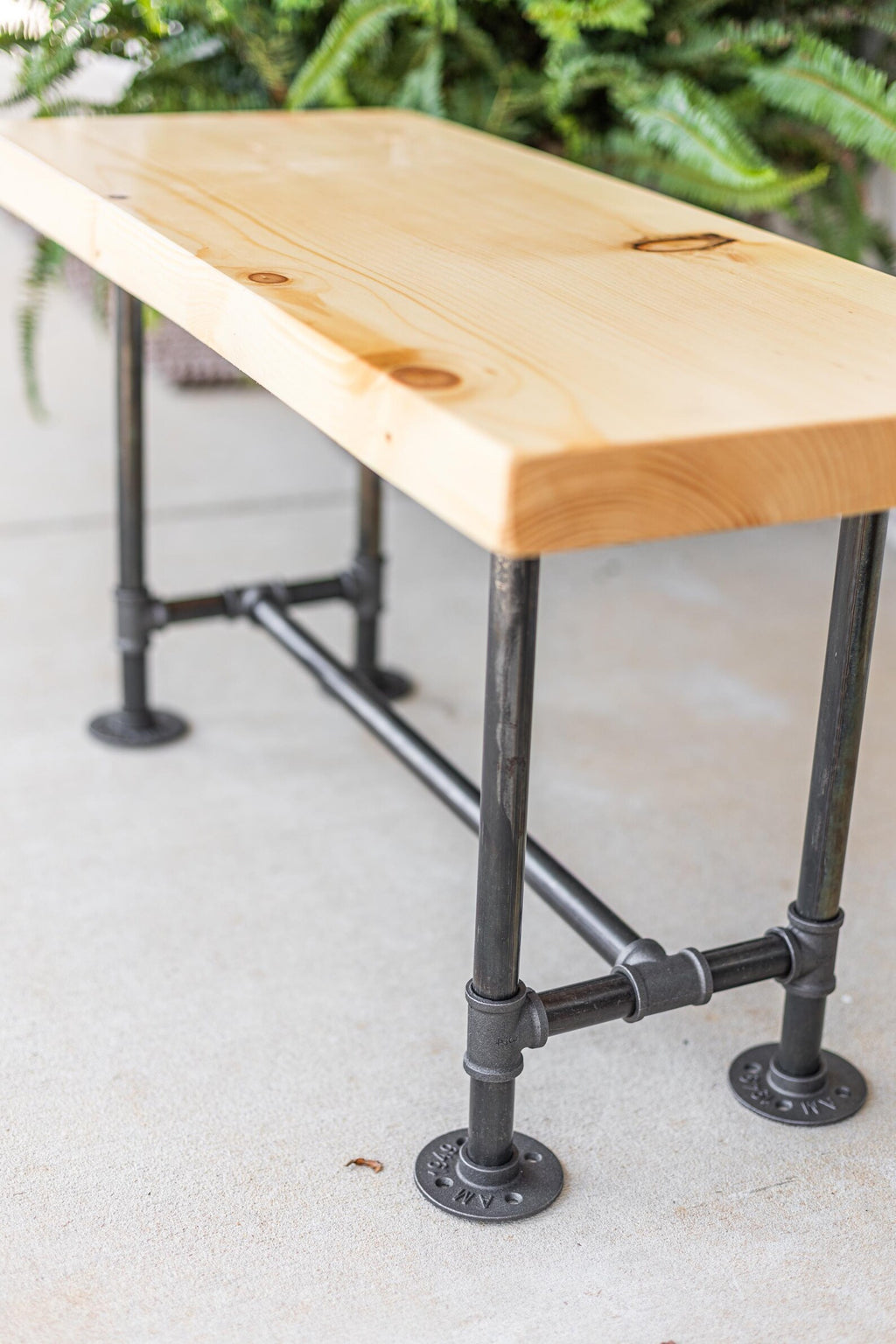May include: A wooden bench with a natural finish and black metal pipe legs. The bench is sitting on a concrete surface.