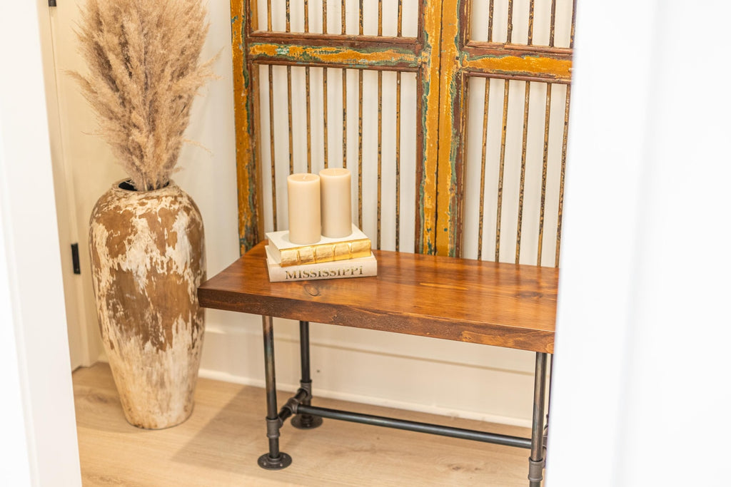 May include: A wooden bench with black metal pipe legs. The bench is in front of a decorative screen with a distressed, yellow paint finish. Two white candles are on top of a stack of books on the bench.