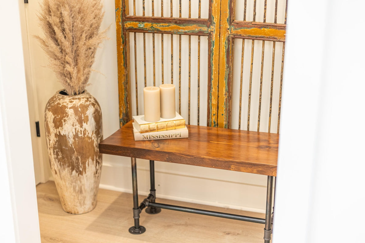 May include: A wooden bench with black metal pipe legs. The bench is in front of a decorative screen with a distressed, yellow paint finish. Two white candles are on top of a stack of books on the bench.