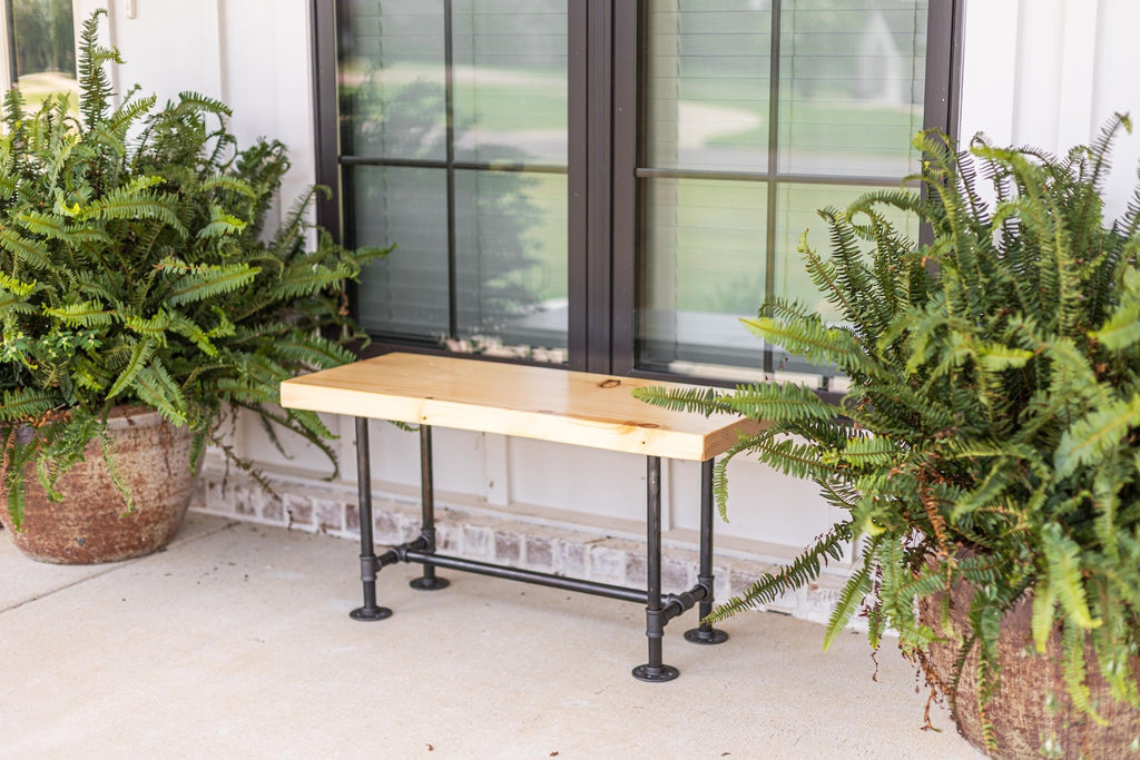 May include: A wooden bench with black metal pipe legs. The bench is in front of a window with black trim and green plants on either side.