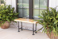 May include: A wooden bench with black metal pipe legs. The bench is in front of a window with black trim and green plants on either side.