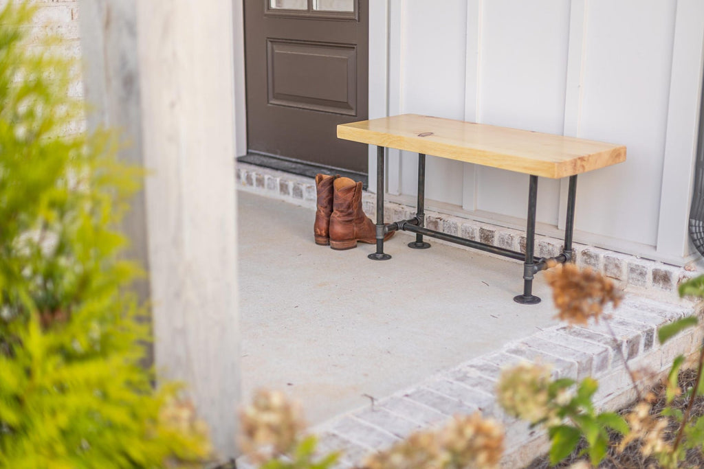 May include: A wooden bench with black metal pipe legs sits on a white brick porch. Two pairs of brown cowboy boots are placed on the porch floor.