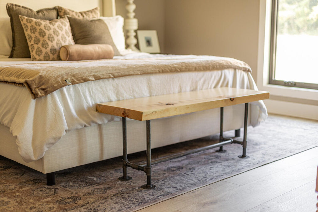 May include: A wooden bench with black metal legs sits in front of a bed with a white duvet and brown throw blanket. The bench has a natural wood finish and the legs are made of industrial pipe.