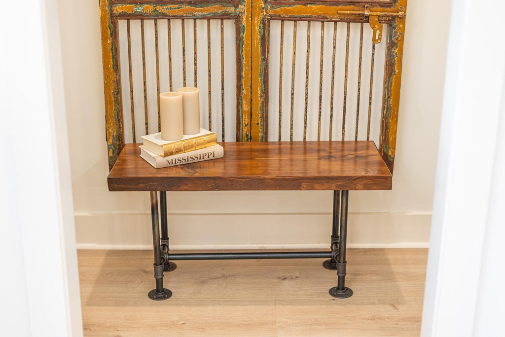 May include: A wooden bench with black metal pipe legs. The bench is sitting in front of a vintage door with a metal grate. Three candles and a stack of books are on top of the bench.