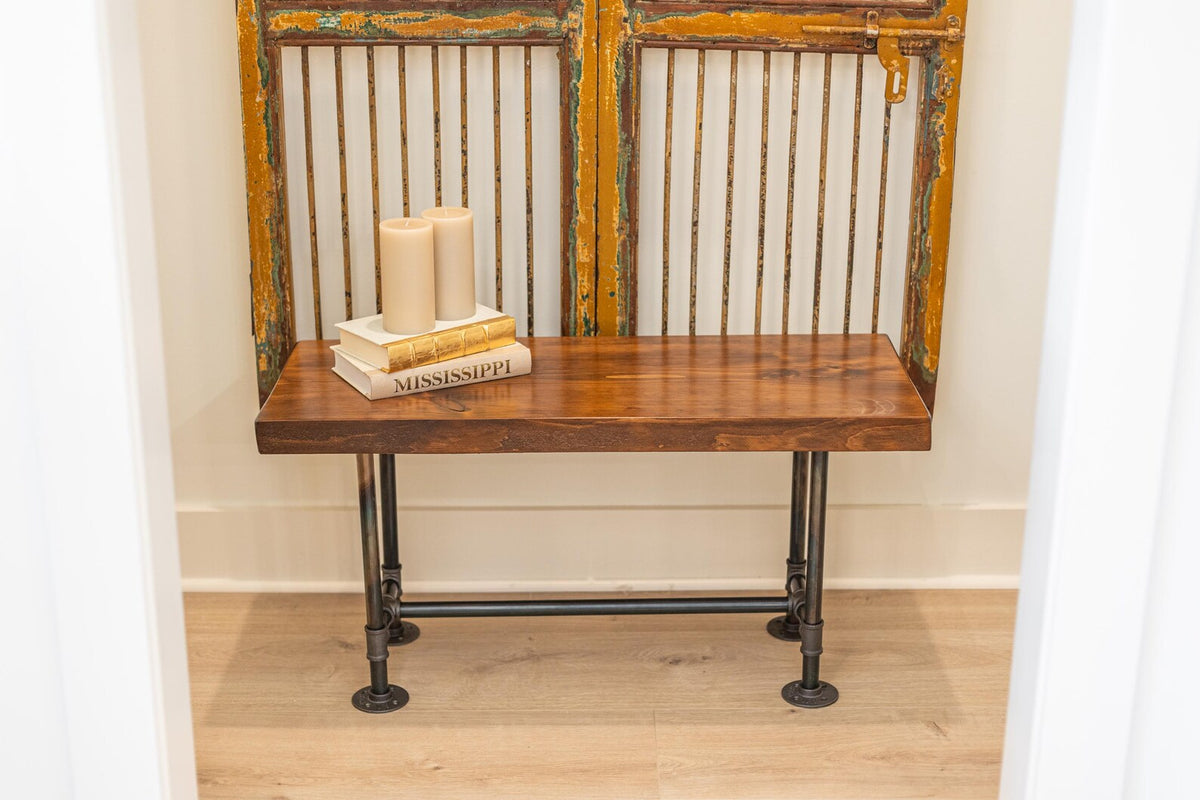 May include: A wooden bench with black metal pipe legs. The bench is sitting in front of a vintage door with a metal grate. Three candles and a stack of books are on top of the bench.