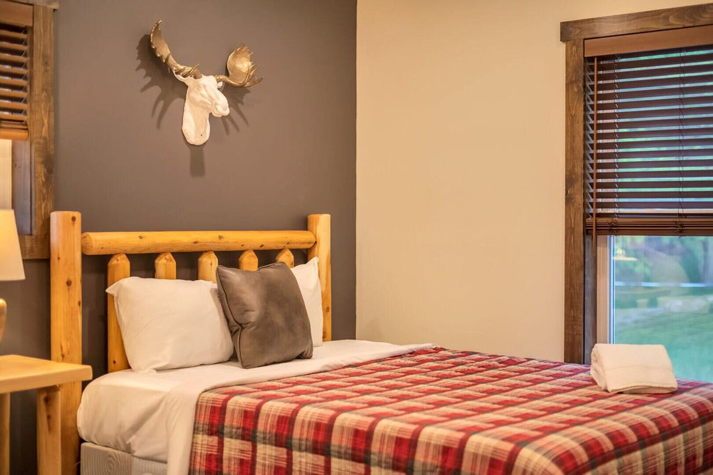 May include: A rustic bedroom scene featuring a bed with a red and black plaid blanket, white pillows, and a gray pillow. The wooden headboard and frame add to the cabin-like aesthetic. A white moose head decorates the wall.