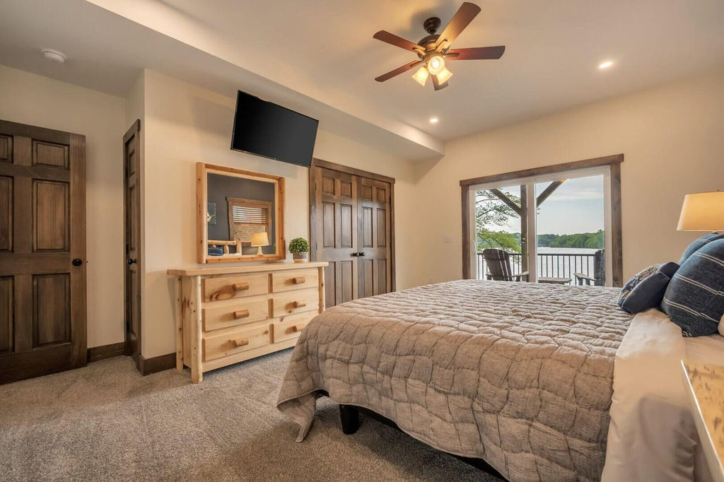May include: A bedroom with a bed, dresser, and TV. The bed has a patterned comforter and pillows. A wooden dresser with six drawers sits below a mirror. A flat-screen TV is mounted on the wall. A ceiling fan and a door are also visible.