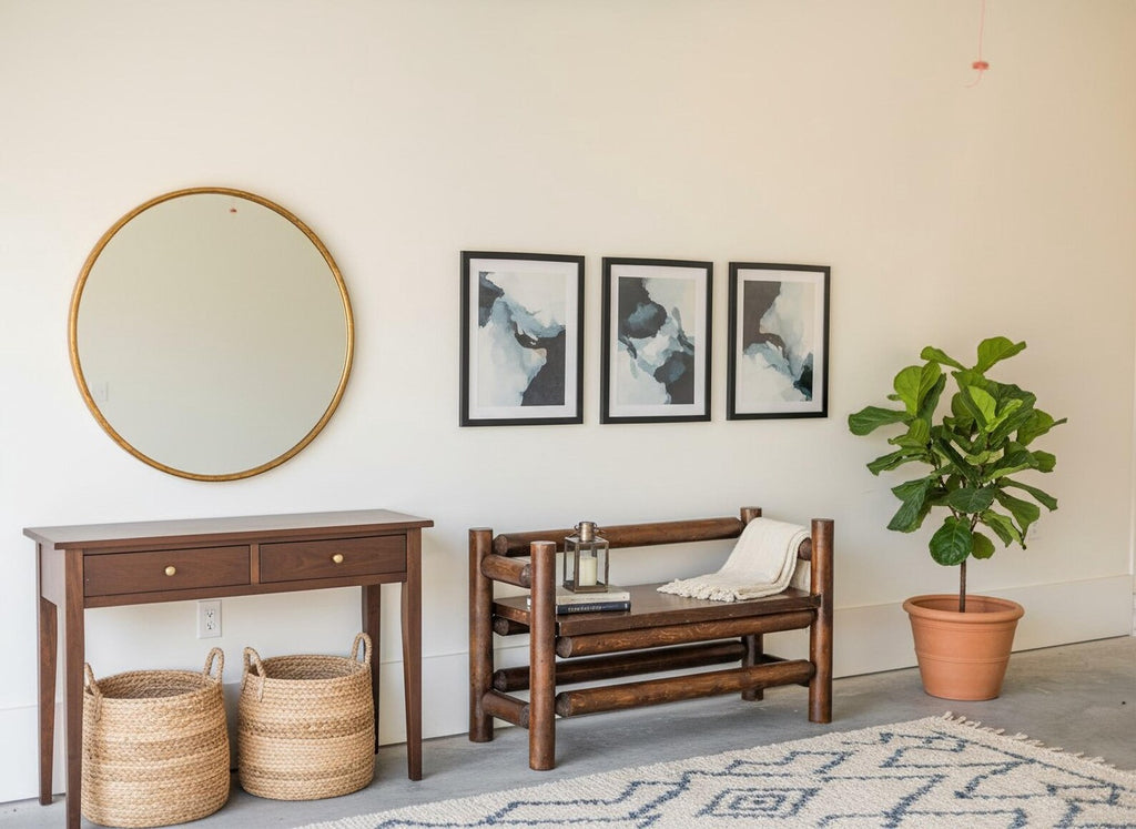 May include: A round gold-framed mirror, a dark wood console table with two drawers, two woven baskets, a dark wood log-style bench with a white throw blanket, three framed abstract prints, and a green plant in a terracotta pot on a rug.