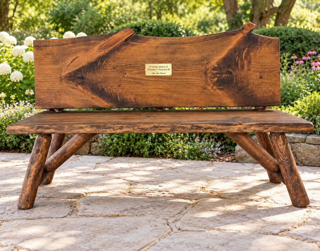 May include: A rustic wooden bench with a live edge backrest and a rectangular seat. The backrest features a centered gold-toned metal plaque with the engraved text: In loving memory of Thomas J. Stewart III, 1946-2022. The bench has four angled log legs.